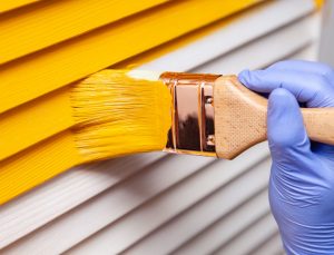 Person applying yellow paint with a paintbrush on the exterior of a house